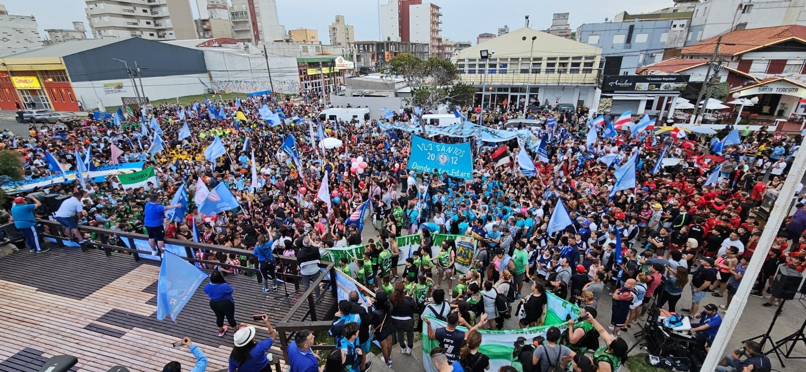 Una gran multitud en una plaza ondeando banderas azules, verdes y rojas. Edificios al fondo.