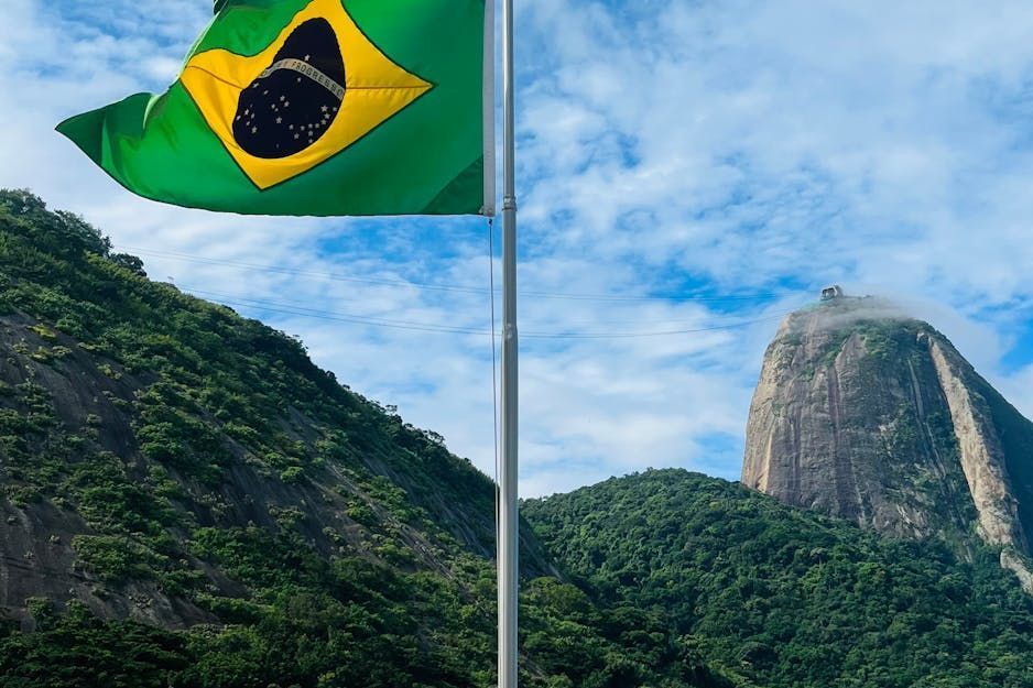 Bandera brasileña ondeando, con montañas verdes y el Pan de Azúcar al fondo.
