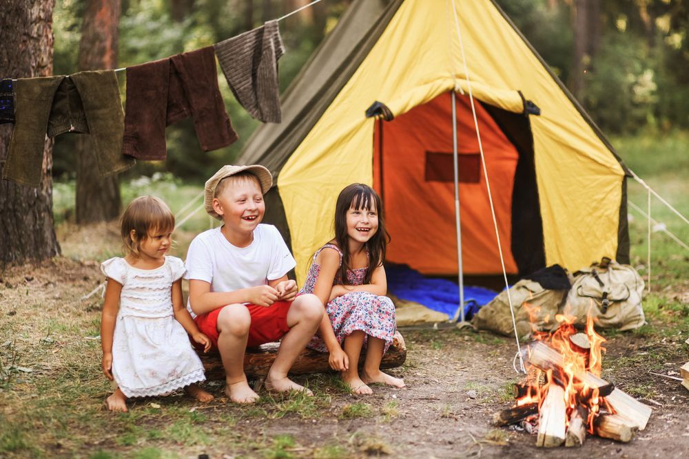 Three children smiling near a campfire and a yellow tent in a forest setting.