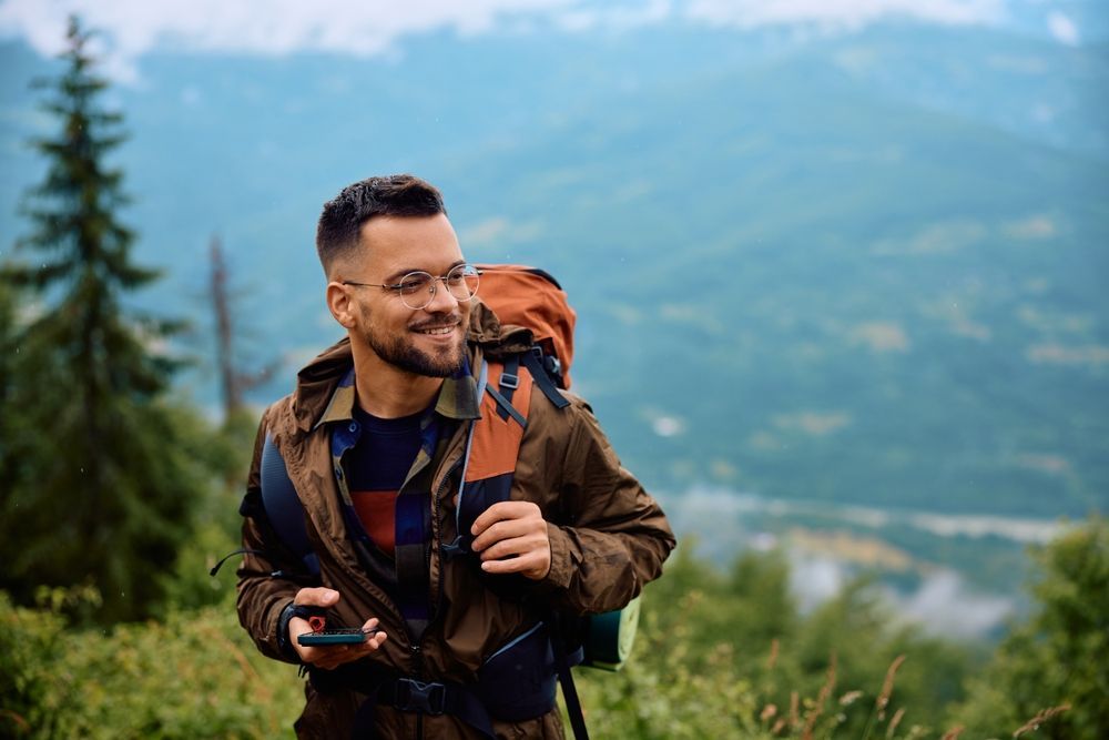 Man with backpack smiles, holding phone, overlooking mountains.