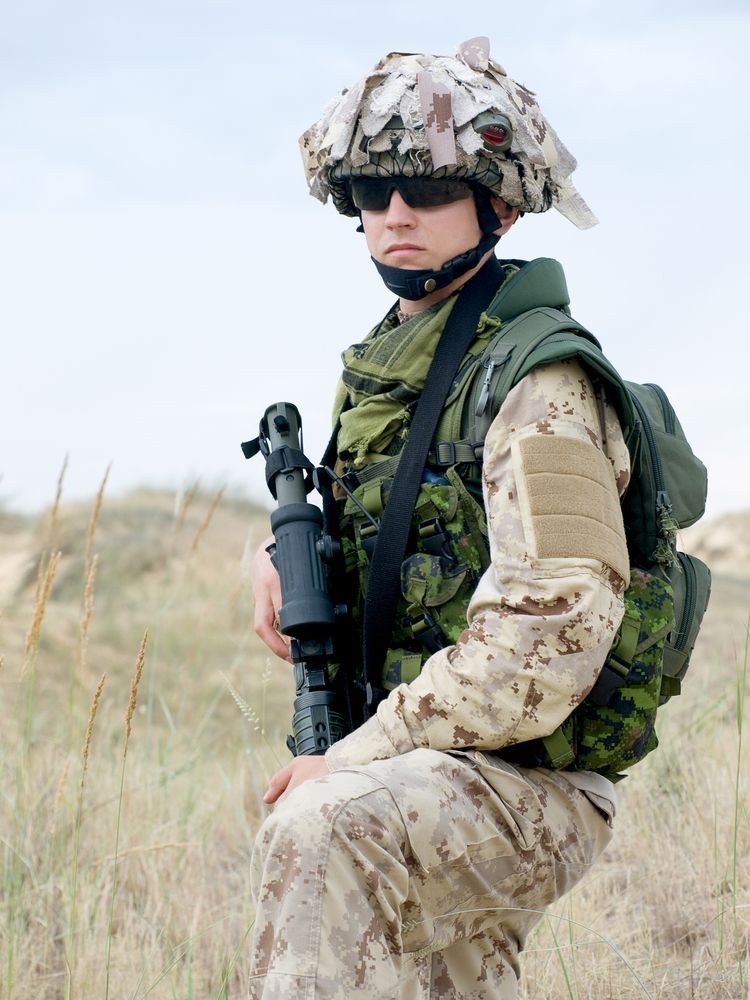 Soldier in desert camouflage, wearing helmet and sunglasses, holding a rifle, standing in a grassy field.