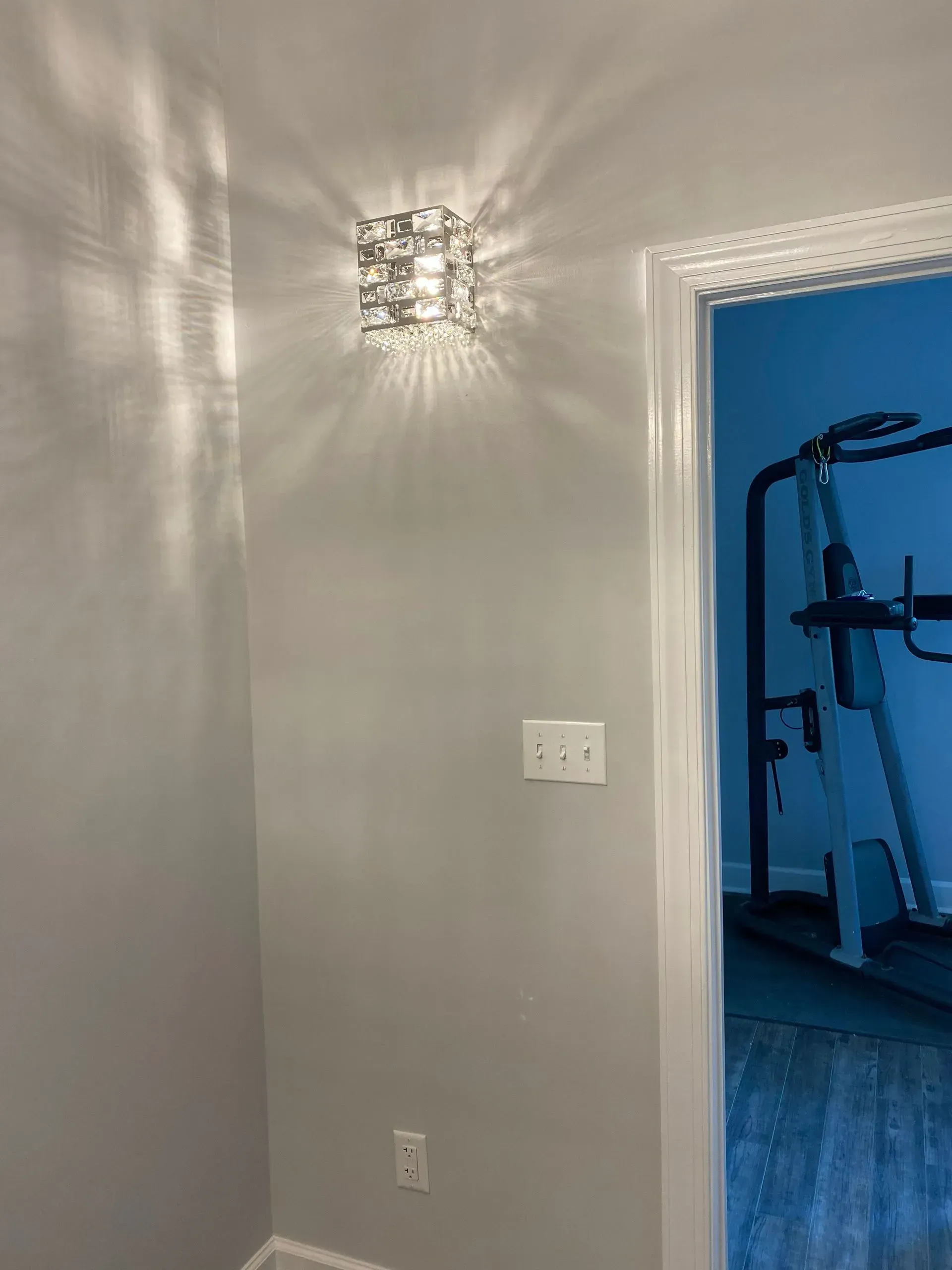 A decorative wall sconce casting light patterns on a gray wall, near a light switch and doorway.