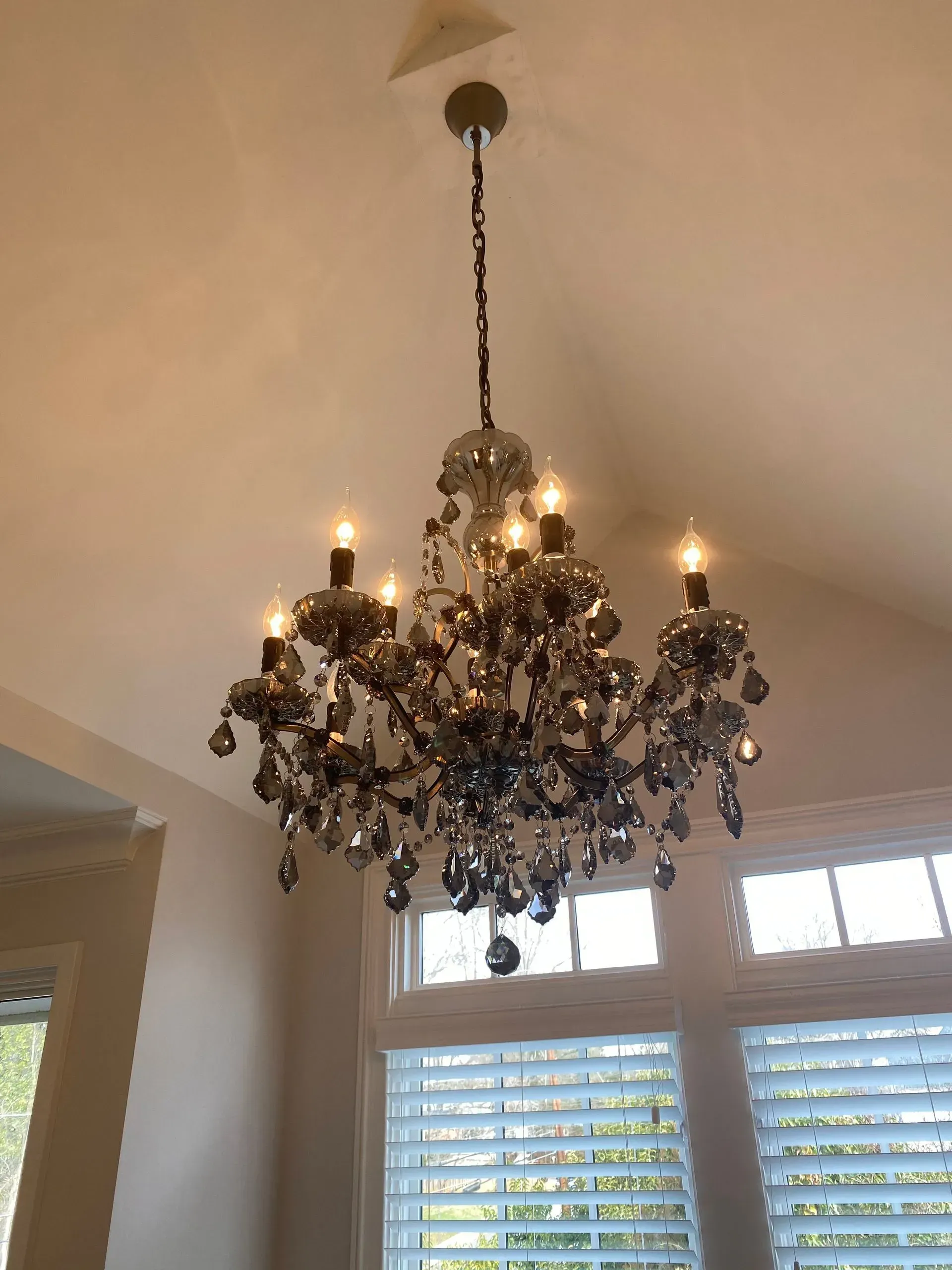 Crystal chandelier hanging from a white ceiling, with lit bulbs and window in the background.