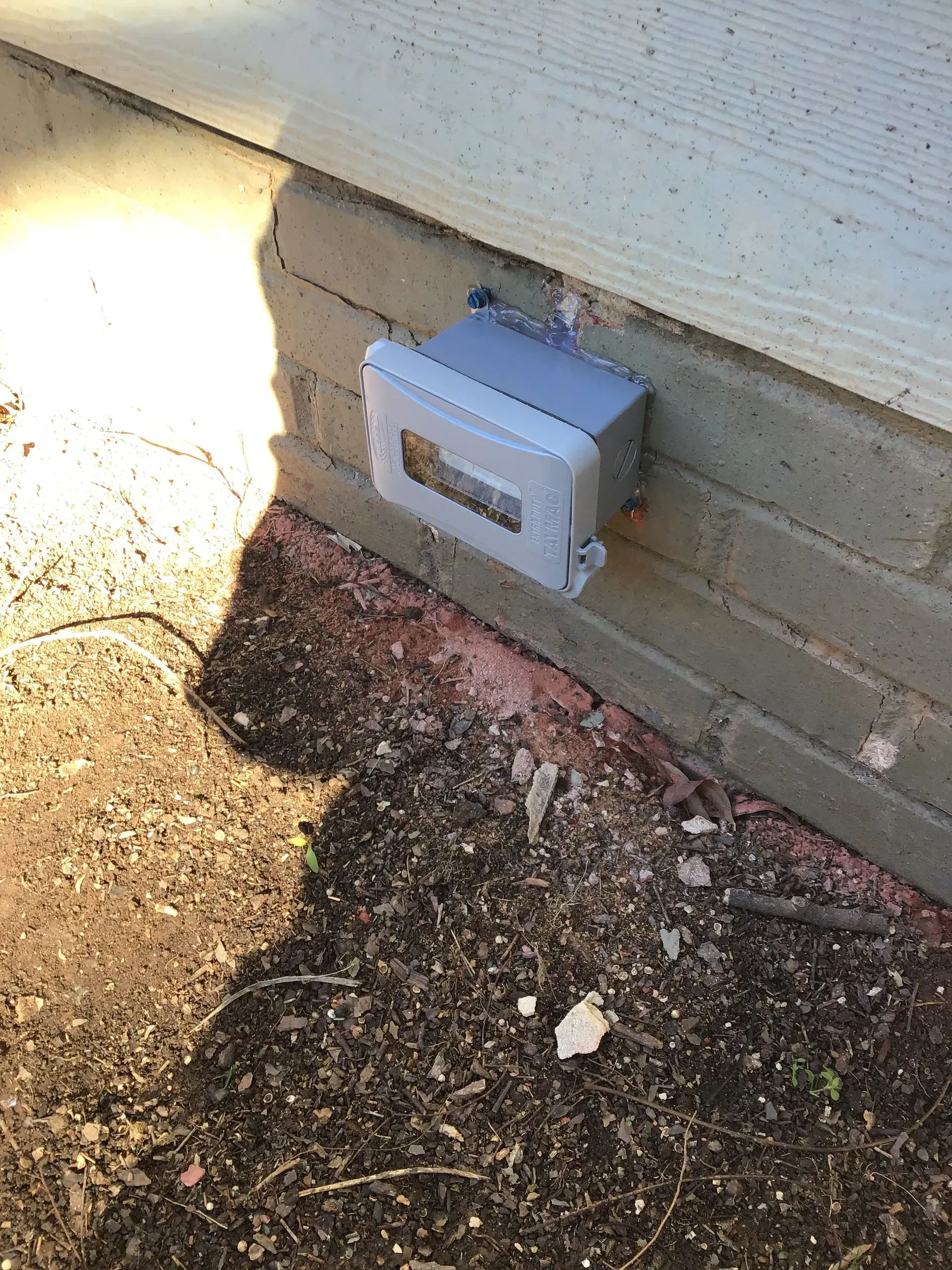 Grey electrical box mounted on brick foundation beneath a building's siding.