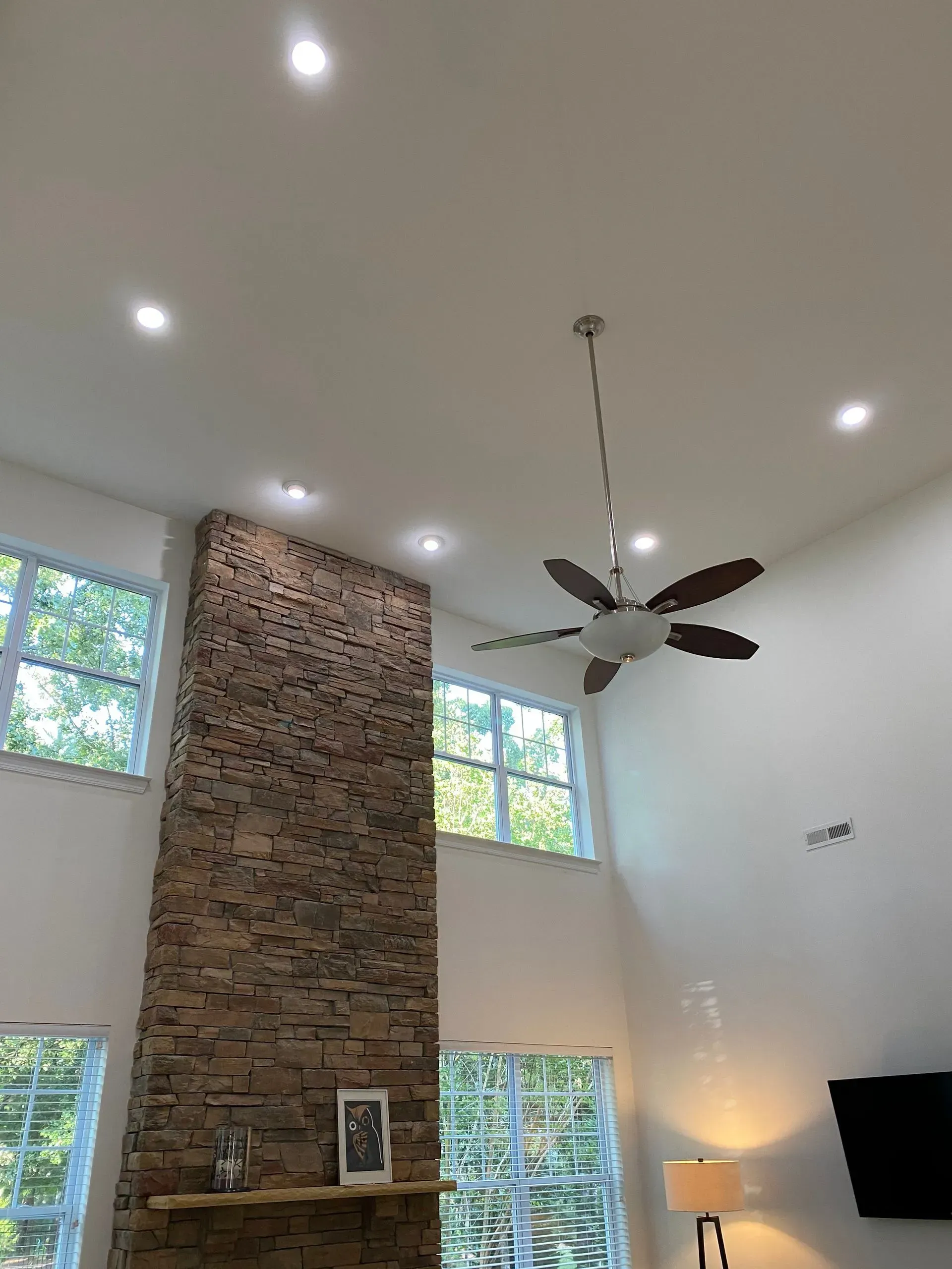 High-ceiling living room with a stone fireplace, windows, recessed lights, and a ceiling fan.
