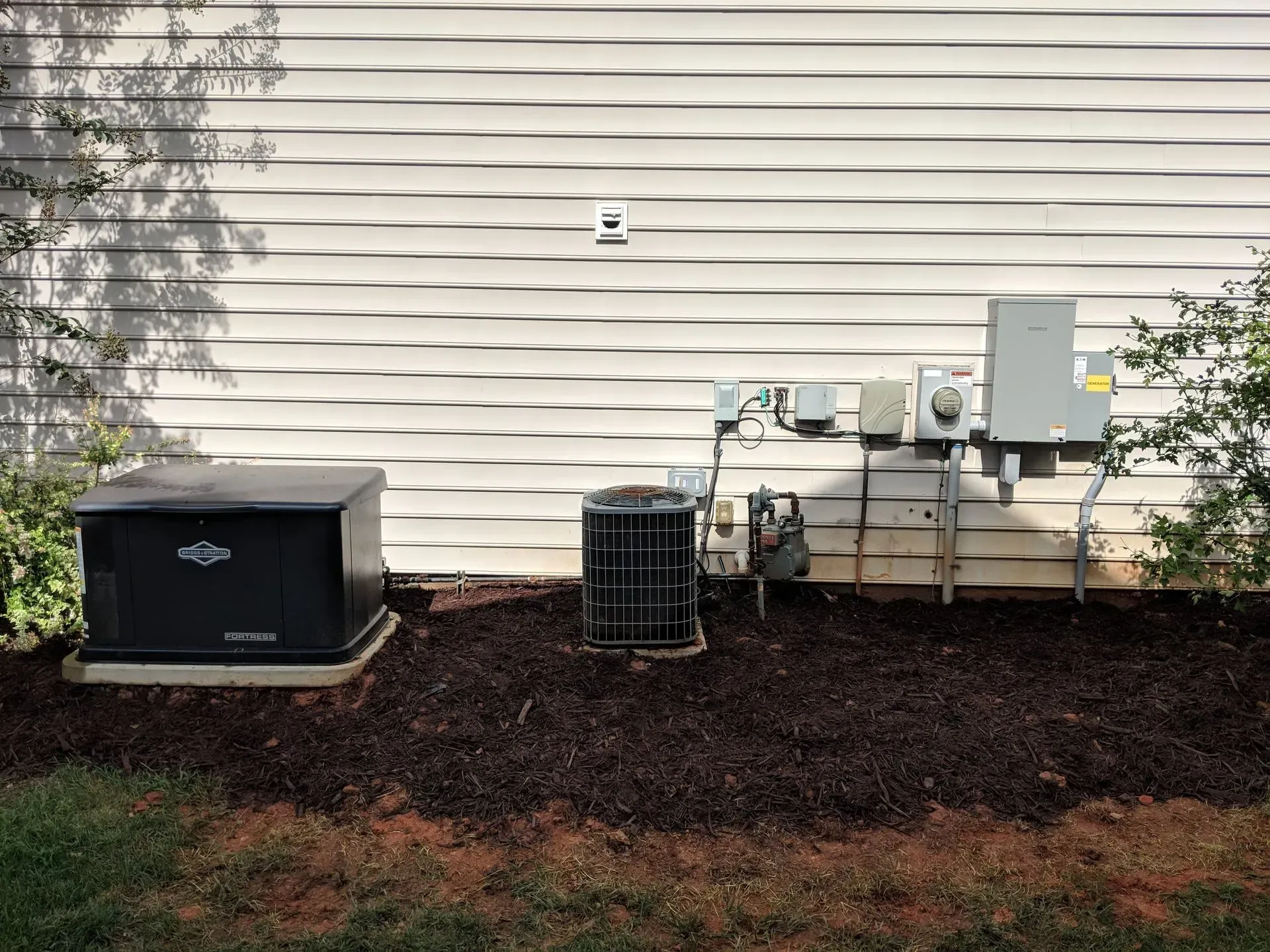 Generator, AC unit, and electrical components installed outside a building on a bed of mulch.
