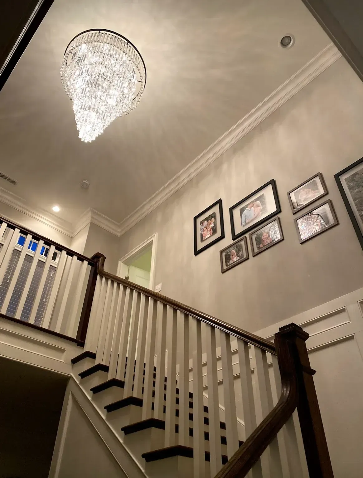 Crystal chandelier hangs above a wooden staircase with white balusters. Family photos on the wall.