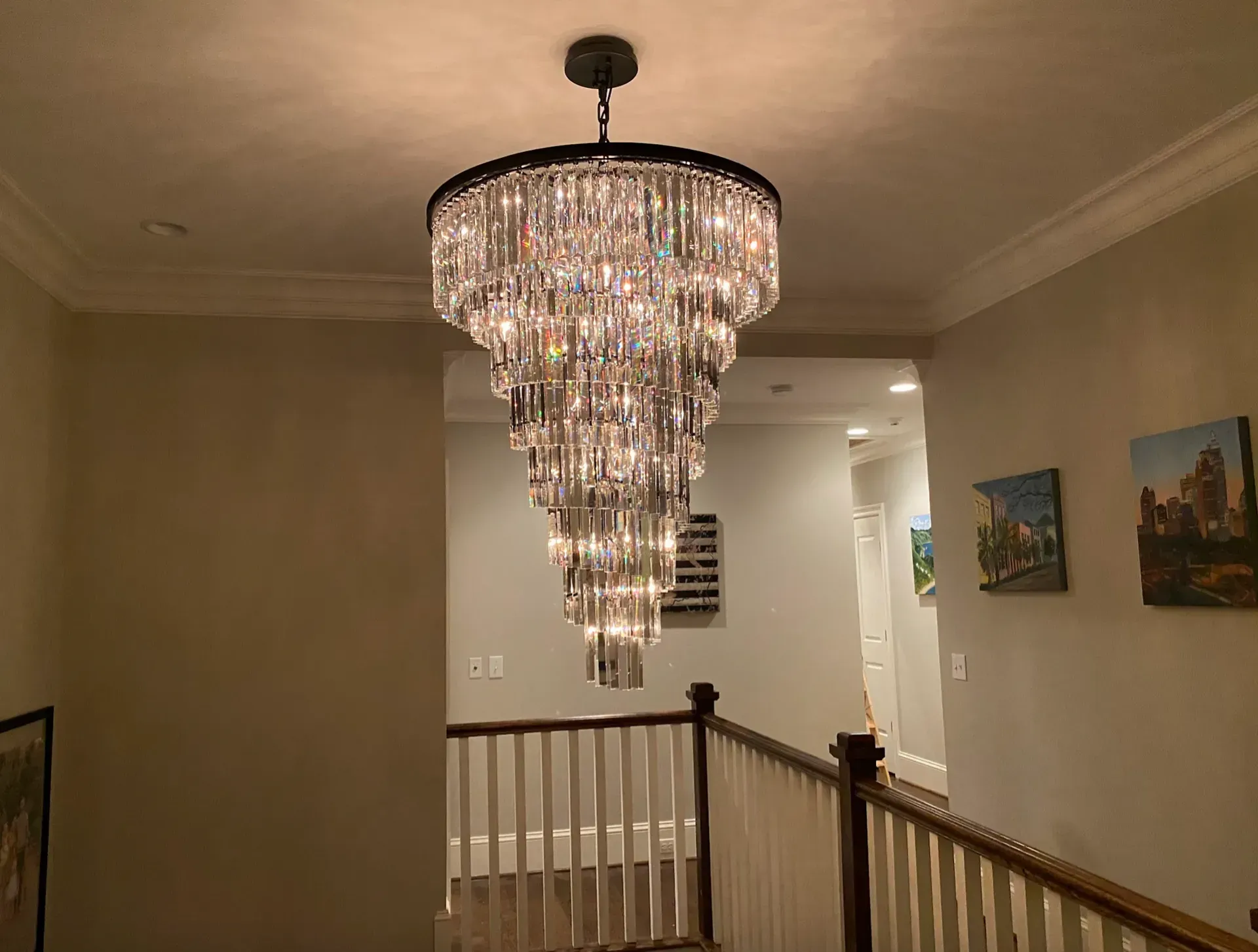 A large, tiered crystal chandelier hangs in a hallway with white walls and a wooden railing.