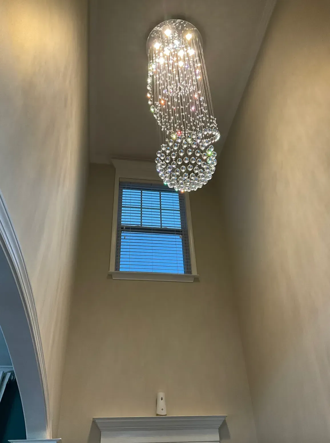 Beaded crystal chandelier hangs in a tall room with a window. Beige walls, white trim.