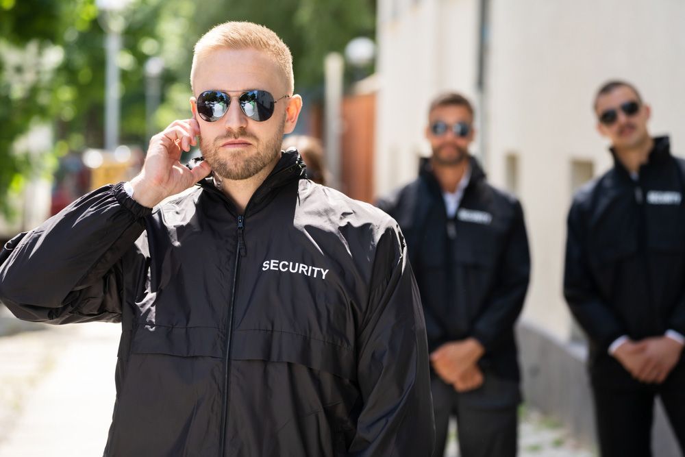 Security Man Wearing Sunglasses And A Security Jacket Talks On A Cell Phone — Professional Security Guards in Chinchilla, QLD