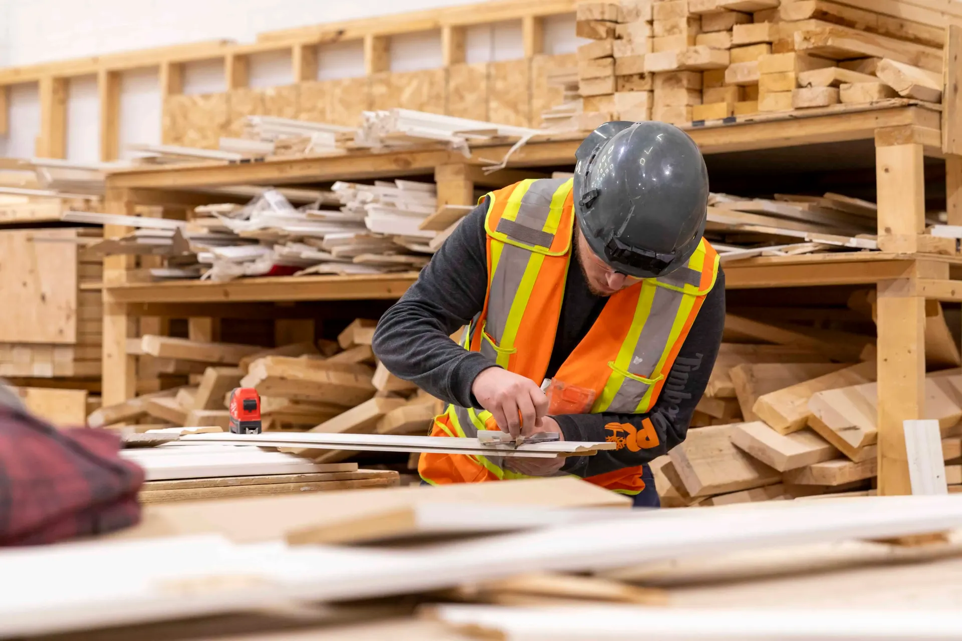 Worker in hard hat and safety vest measuring wood in a lumber warehouse