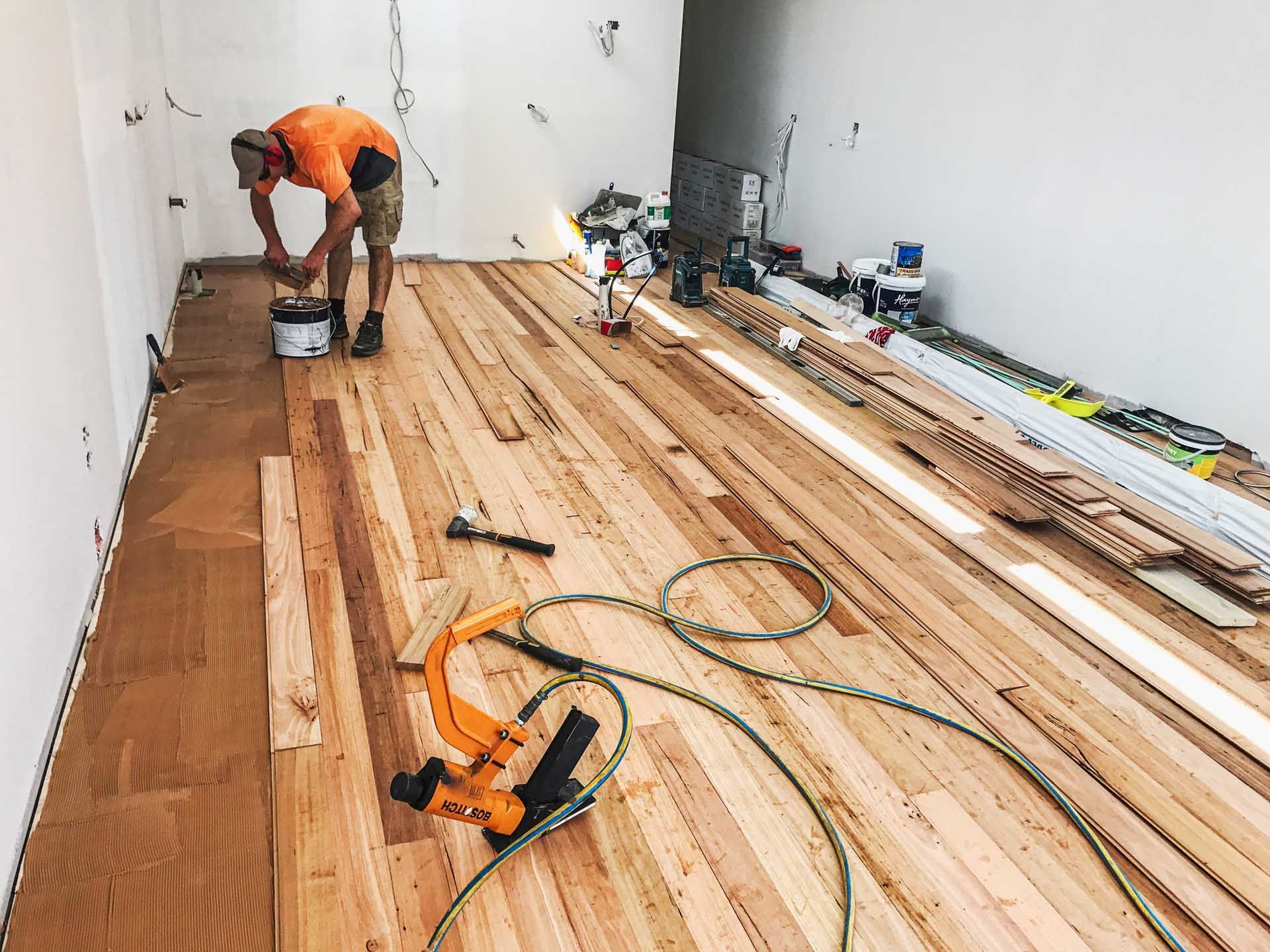 Worker installing wooden floorboards in a bright, empty room with tools and materials scattered nearby.