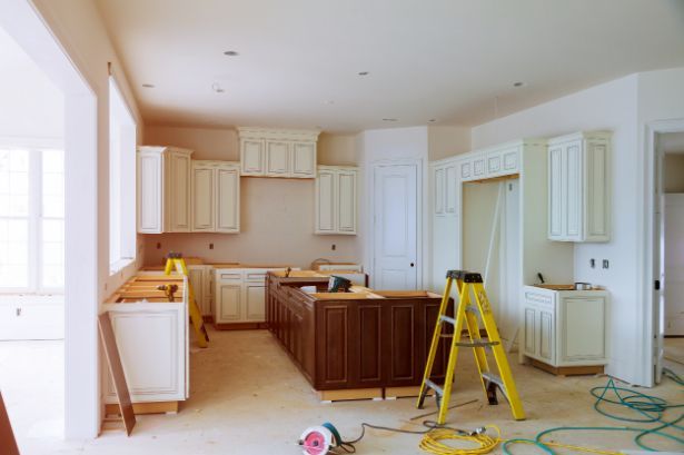 Kitchen renovation in progress: cream cabinets, dark brown island, yellow ladder, tools, and unfinished floor.