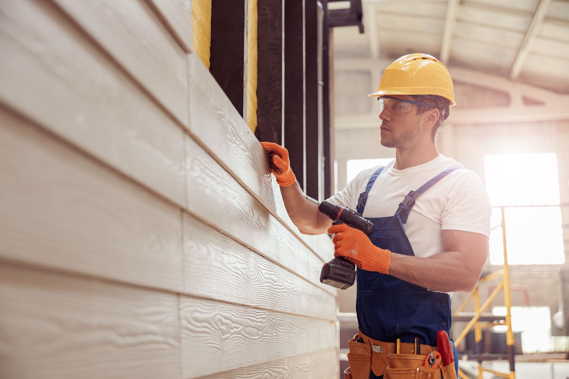Construction worker in yellow hard hat and blue overalls painting a wall with a spray gun.