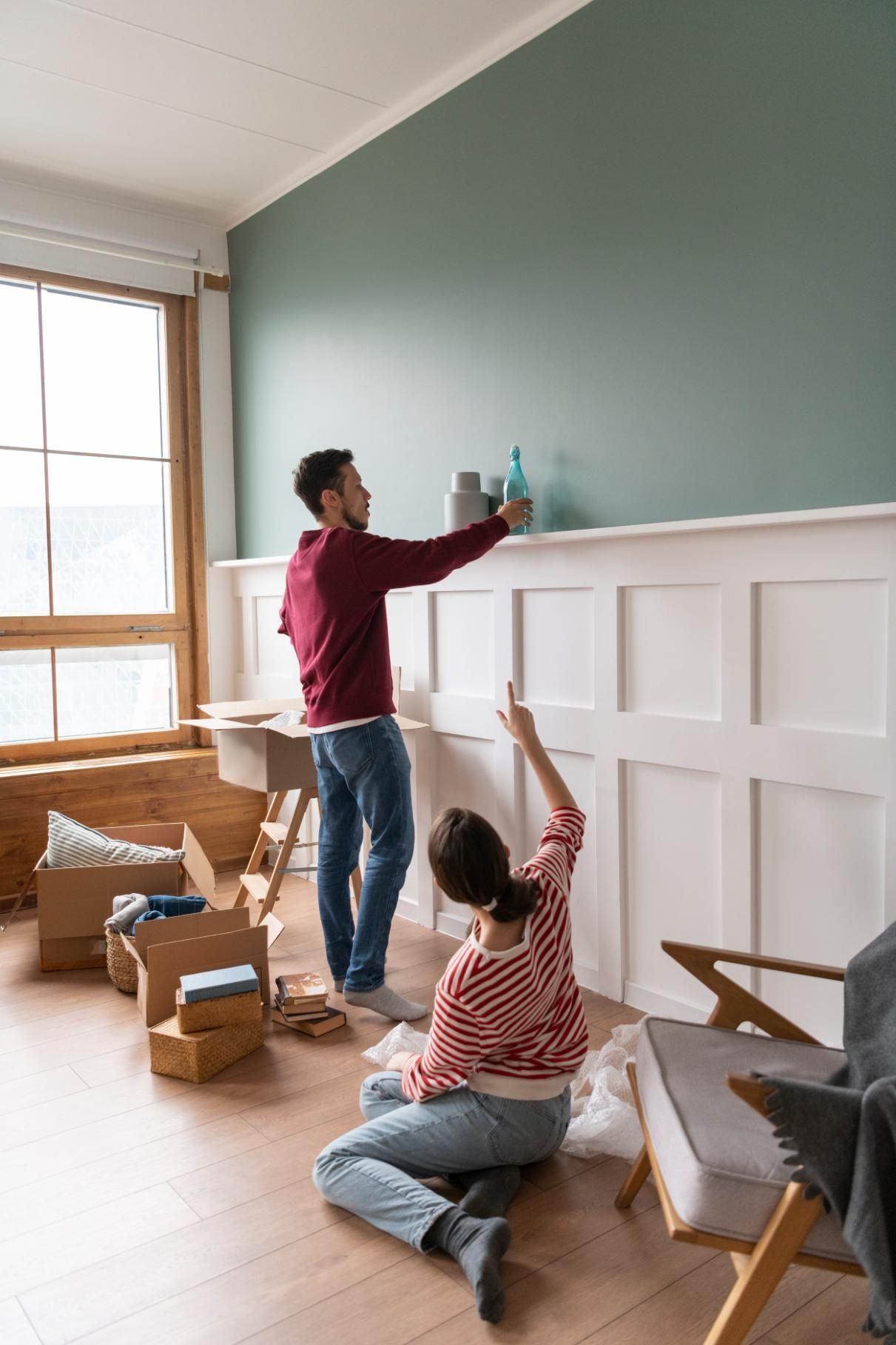 Couple decorates a room with boxes and furniture. Man reaches for decor on a shelf, woman points upwards.