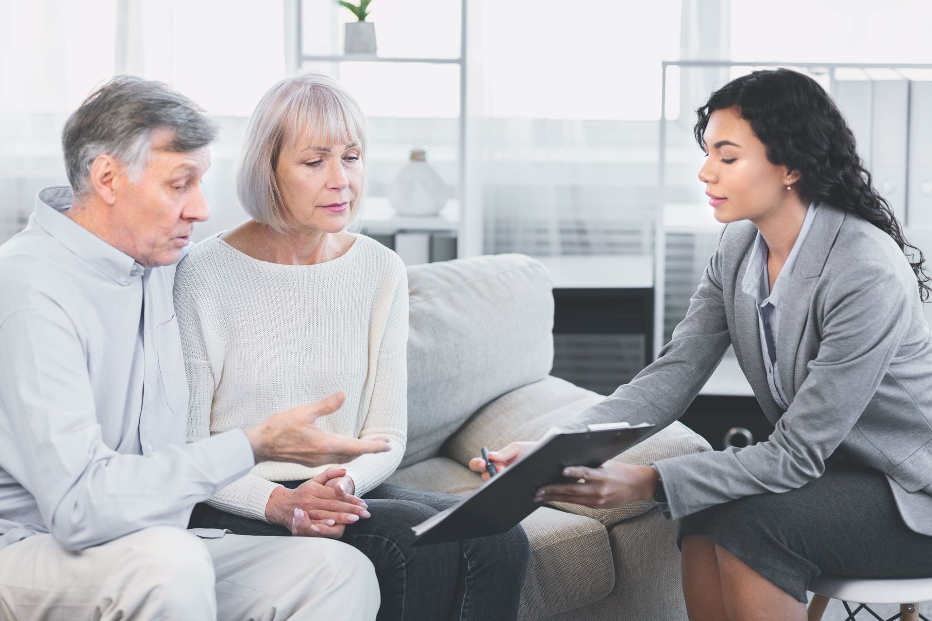 A person in a suit reviews paperwork with a couple on a couch; indoor setting.