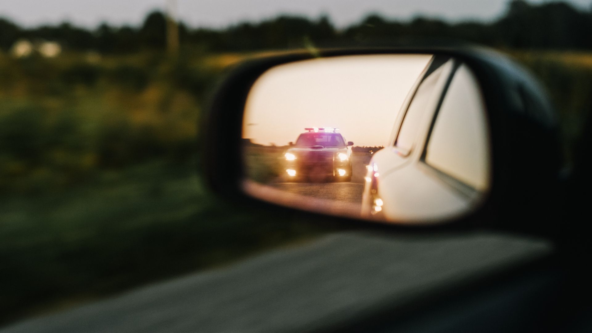 Car's side mirror reflecting a police car with flashing lights on a road at dusk.