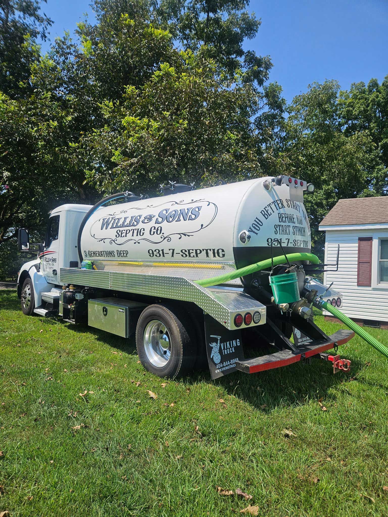 White septic service truck on grass beside a house, with green equipment and tree-lined yard.