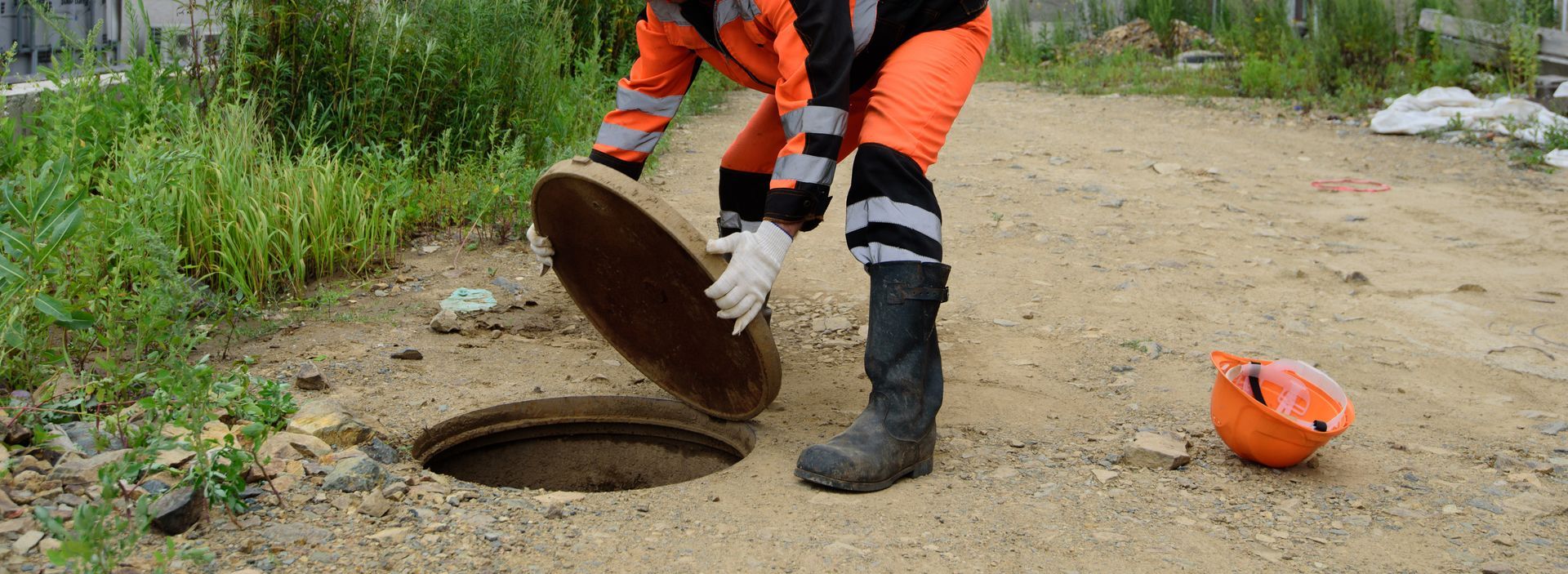 Worker in orange safety gear lifting a manhole cover on a dirt road beside grass and an orange helmet. Worker in orange safety gear lifting a manhole cover on a dirt road beside grass and an orange helmet.