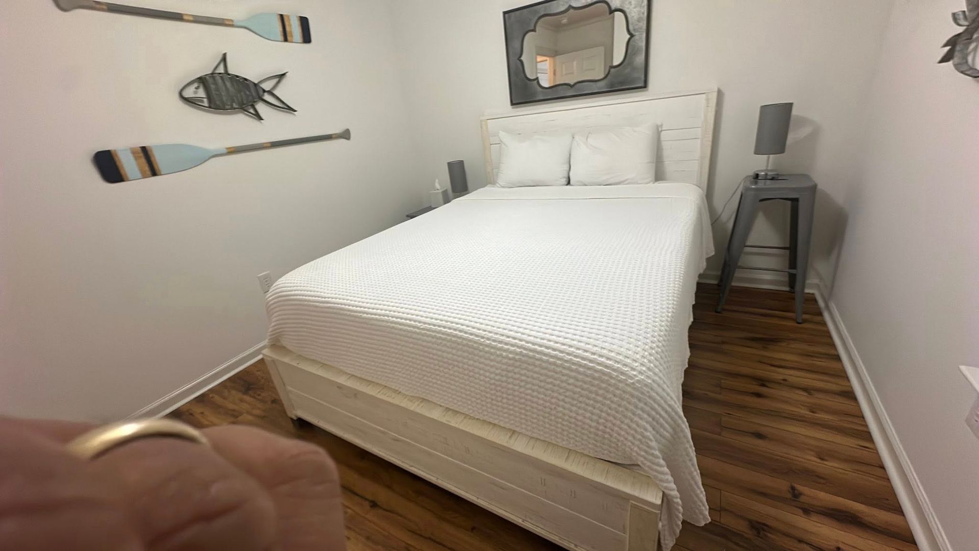 A white bed in a room with light wood floors, nautical-themed wall decor, and a small grey bedside table with a lamp.