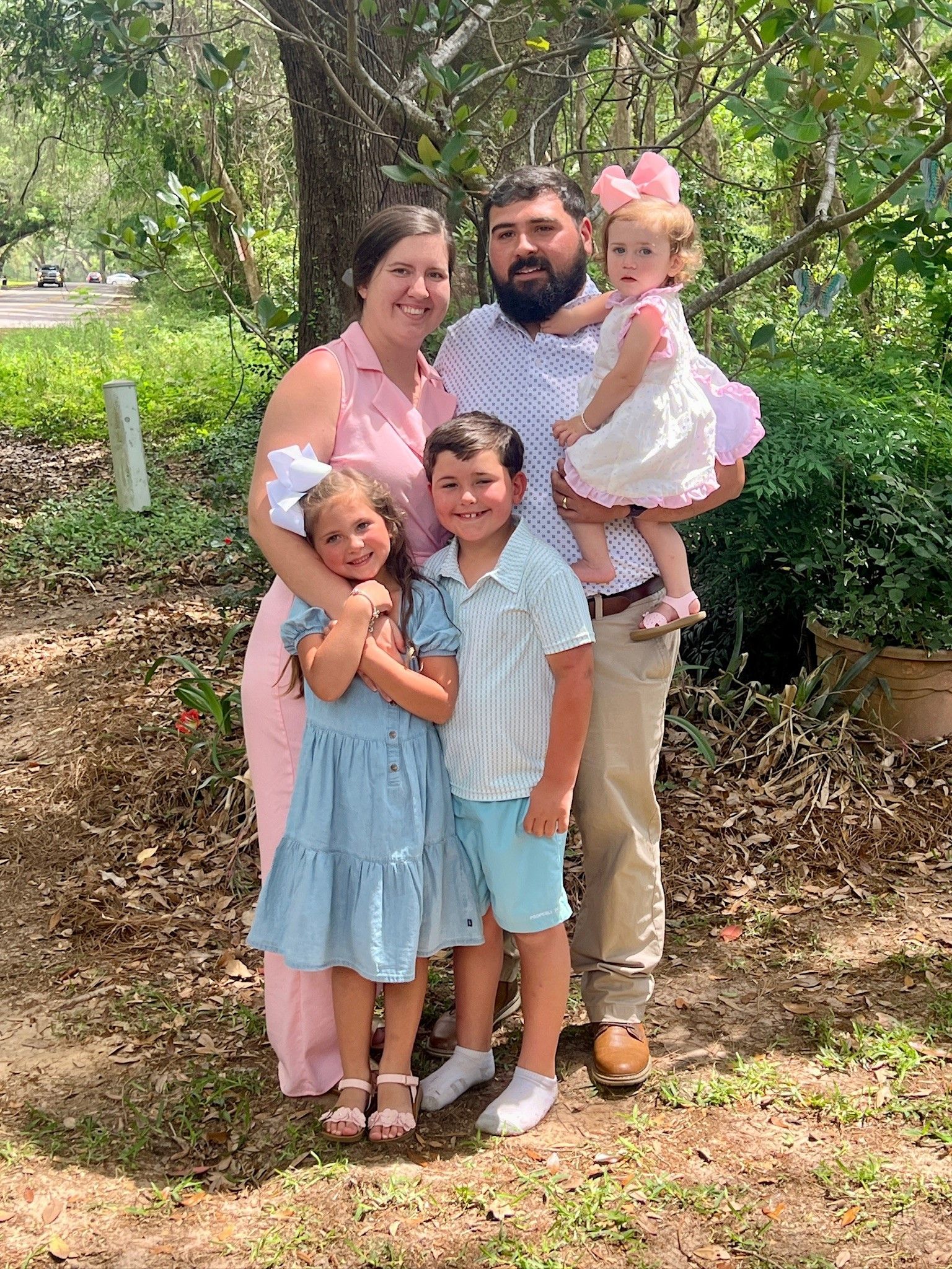 A family of five poses outdoors in a wooded area, smiling for the camera while dressed in coordinated pastel outfits.