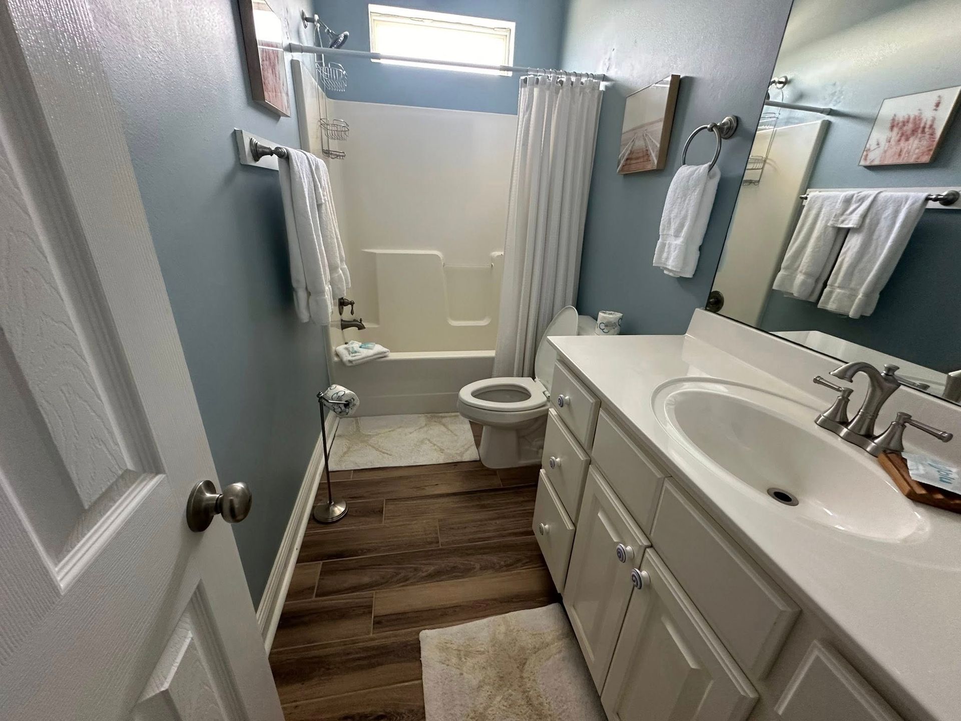 A small bathroom with a white vanity, a tub-shower combo, blue walls, wood-look flooring, and white towels.