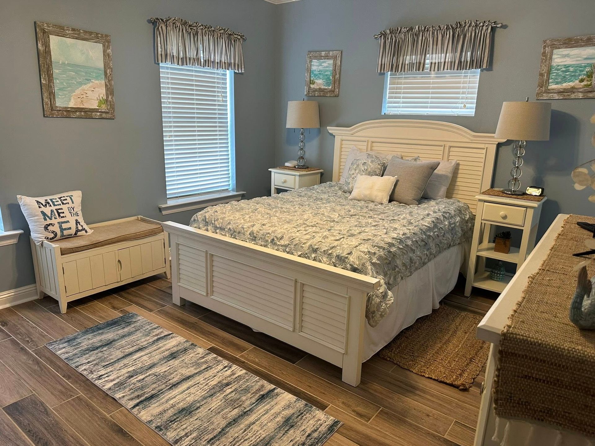 A coastal-themed bedroom with blue walls, a white wood bed, two nightstands, a storage bench, and beach-themed artwork.