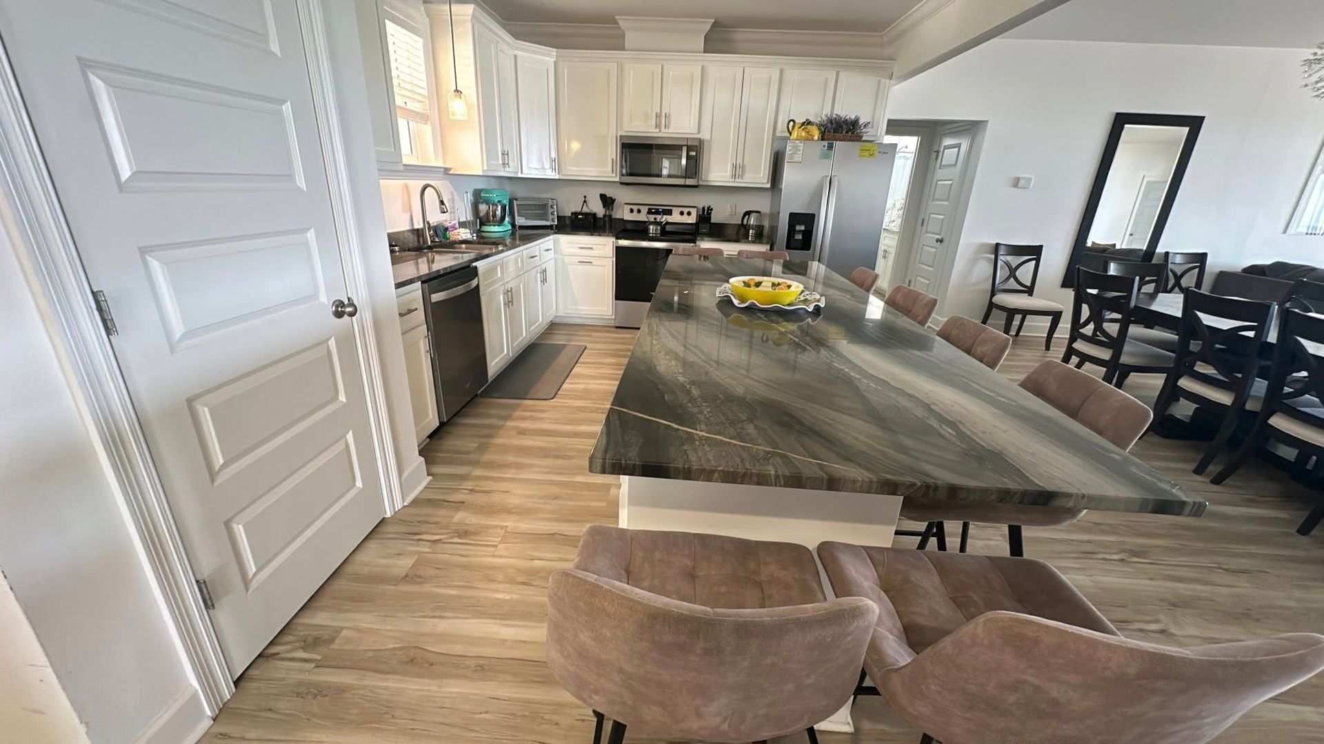 A modern kitchen with white cabinets, dark stone countertops, a large island with three chairs, and a dining area nearby.