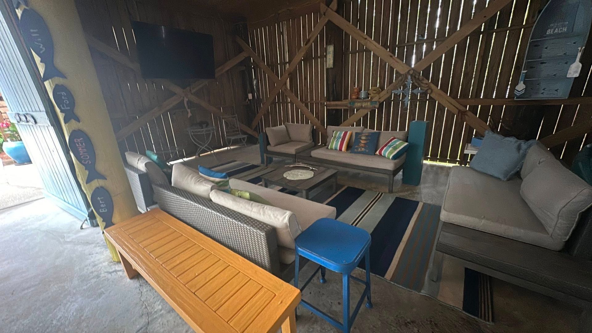 A covered outdoor patio with neutral-toned sofas, a wooden coffee table, a yellow bench, and a blue stool on a rug.