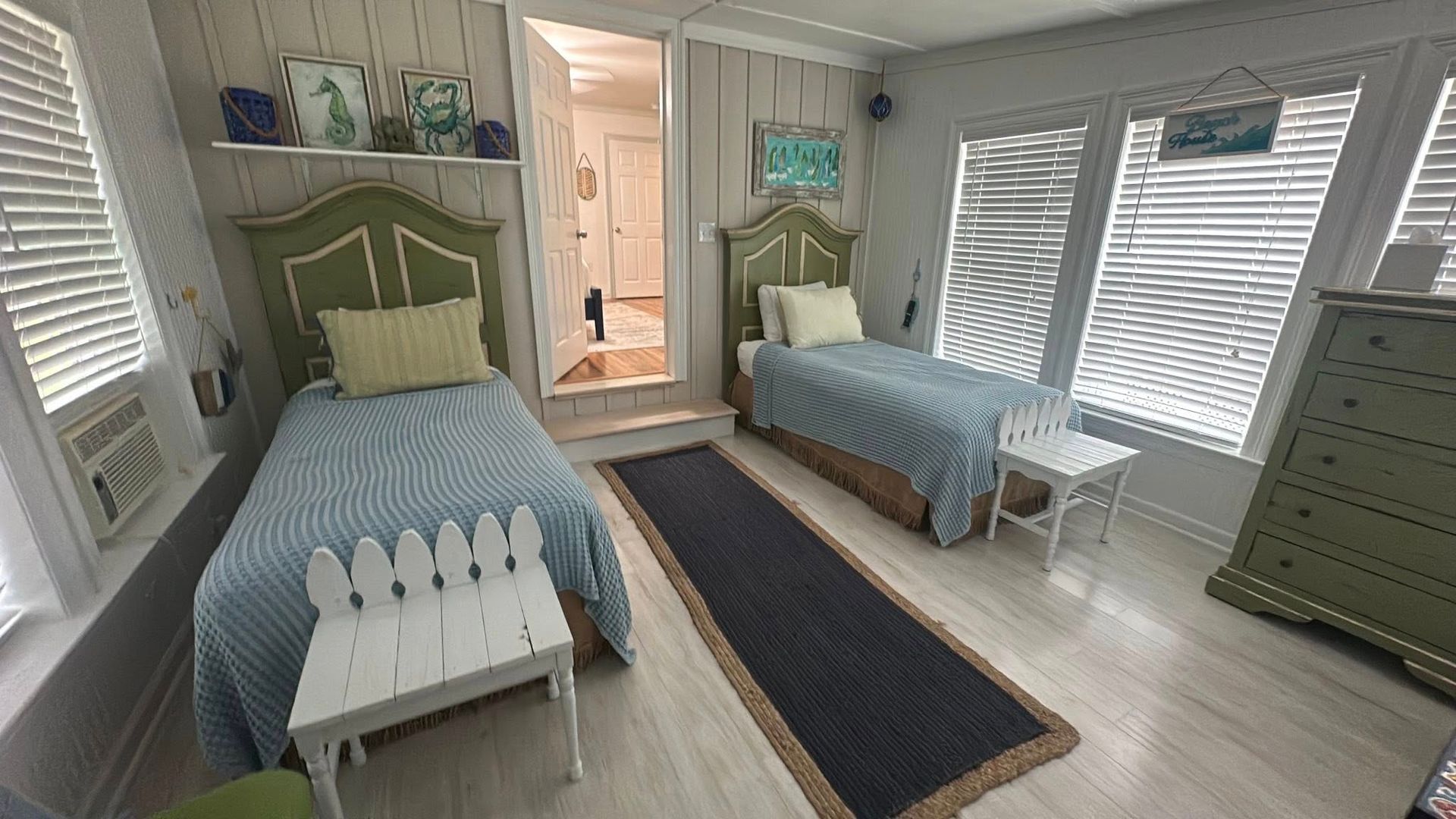 Two twin beds with green headboards and blue patterned quilts in a room with light wood walls and a dark runner rug.