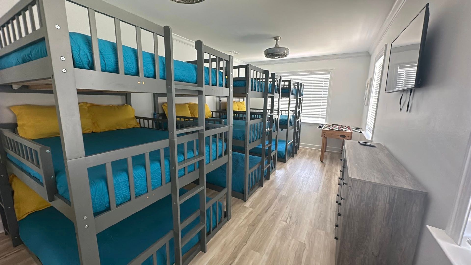 A room featuring rows of grey bunk beds with blue bedding and yellow pillows, a wooden side table, and a wall-mounted TV.
