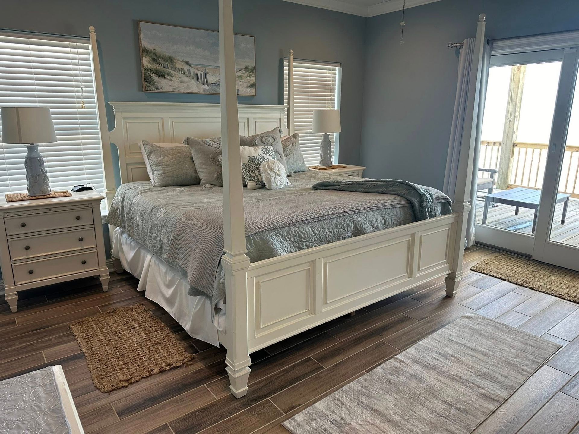 A bedroom with a white four-poster bed, wood floors, two bedside tables with lamps, and a view to a balcony.