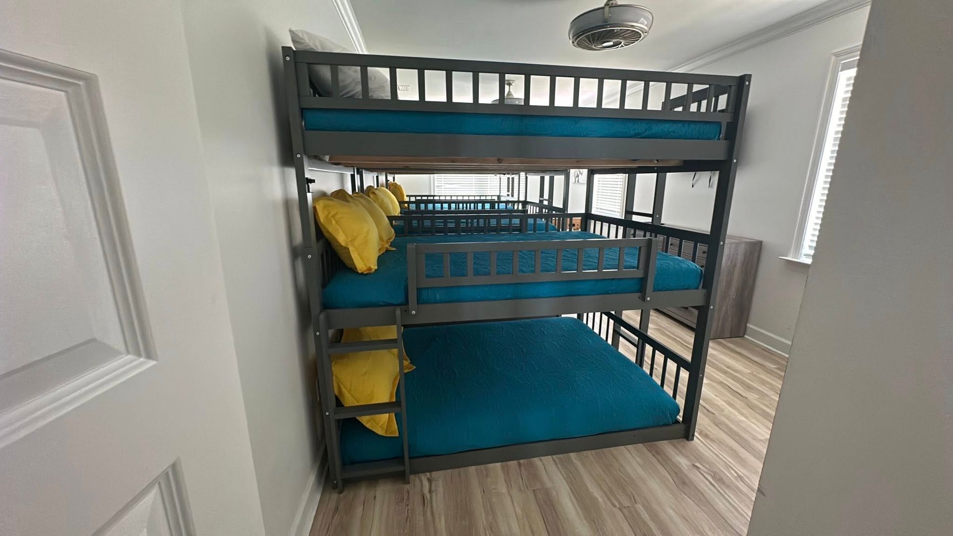 A dark gray triple-bunk bed with teal bedding and yellow pillows in a bright room with light wood-style flooring.