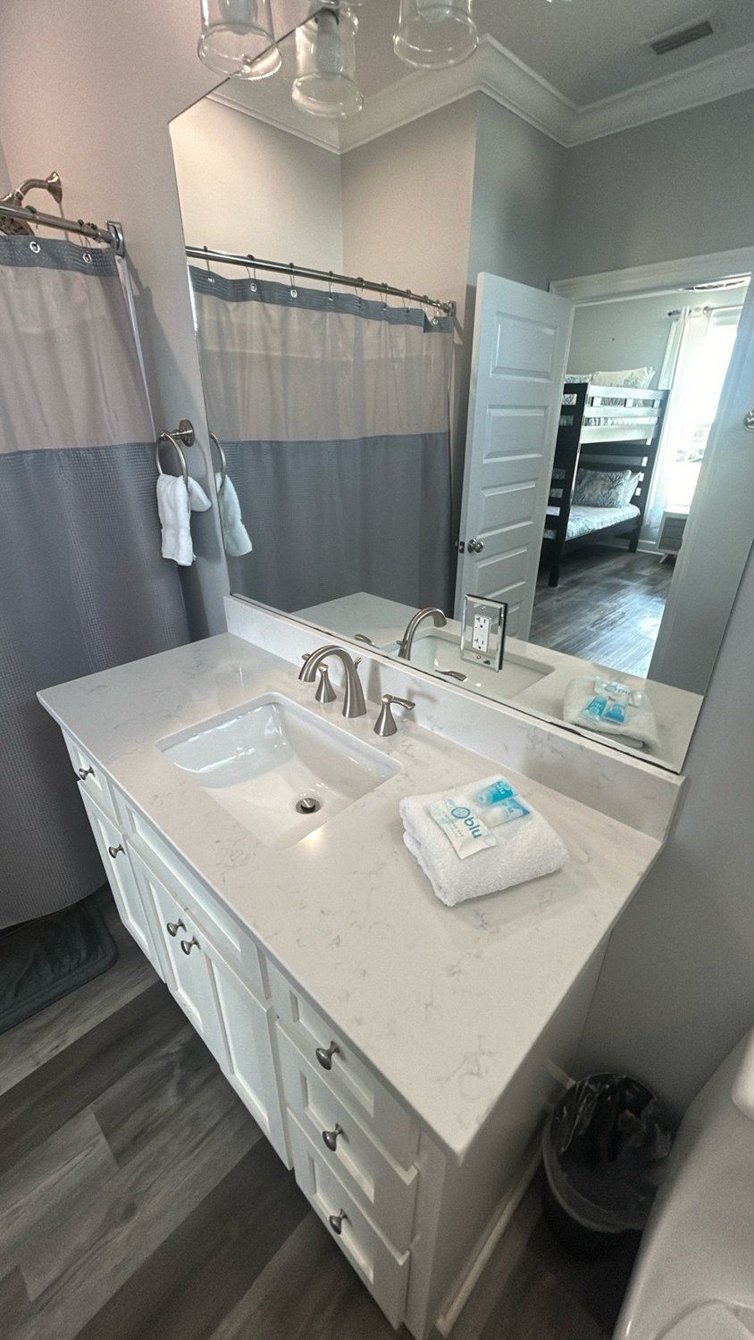 A modern bathroom with a white vanity, quartz countertop, gray shower curtain, and a mirror reflecting a bunk bed.