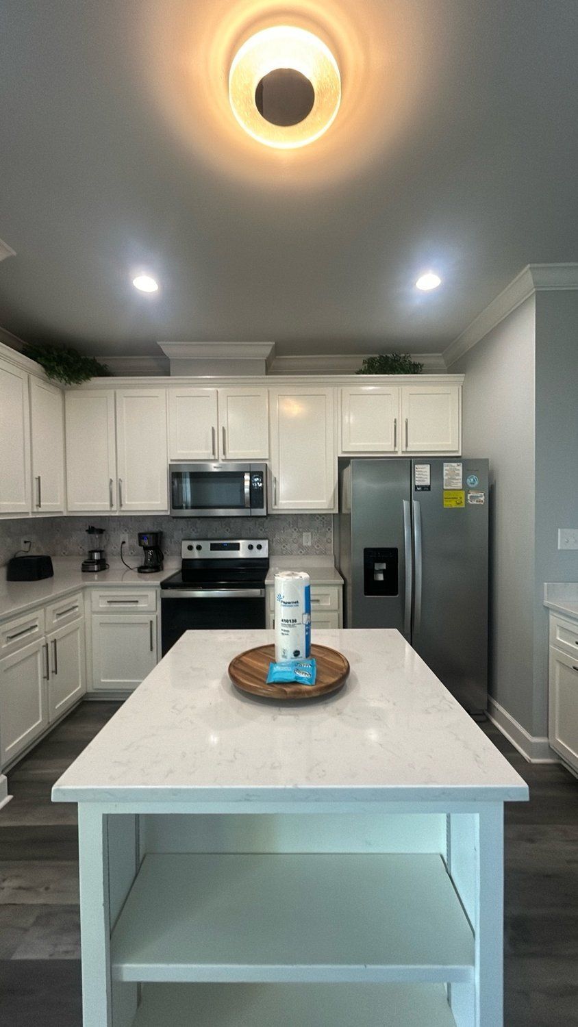 A white kitchen featuring a central island with a wooden tray, stainless steel appliances, and cabinets under warm lighting.
