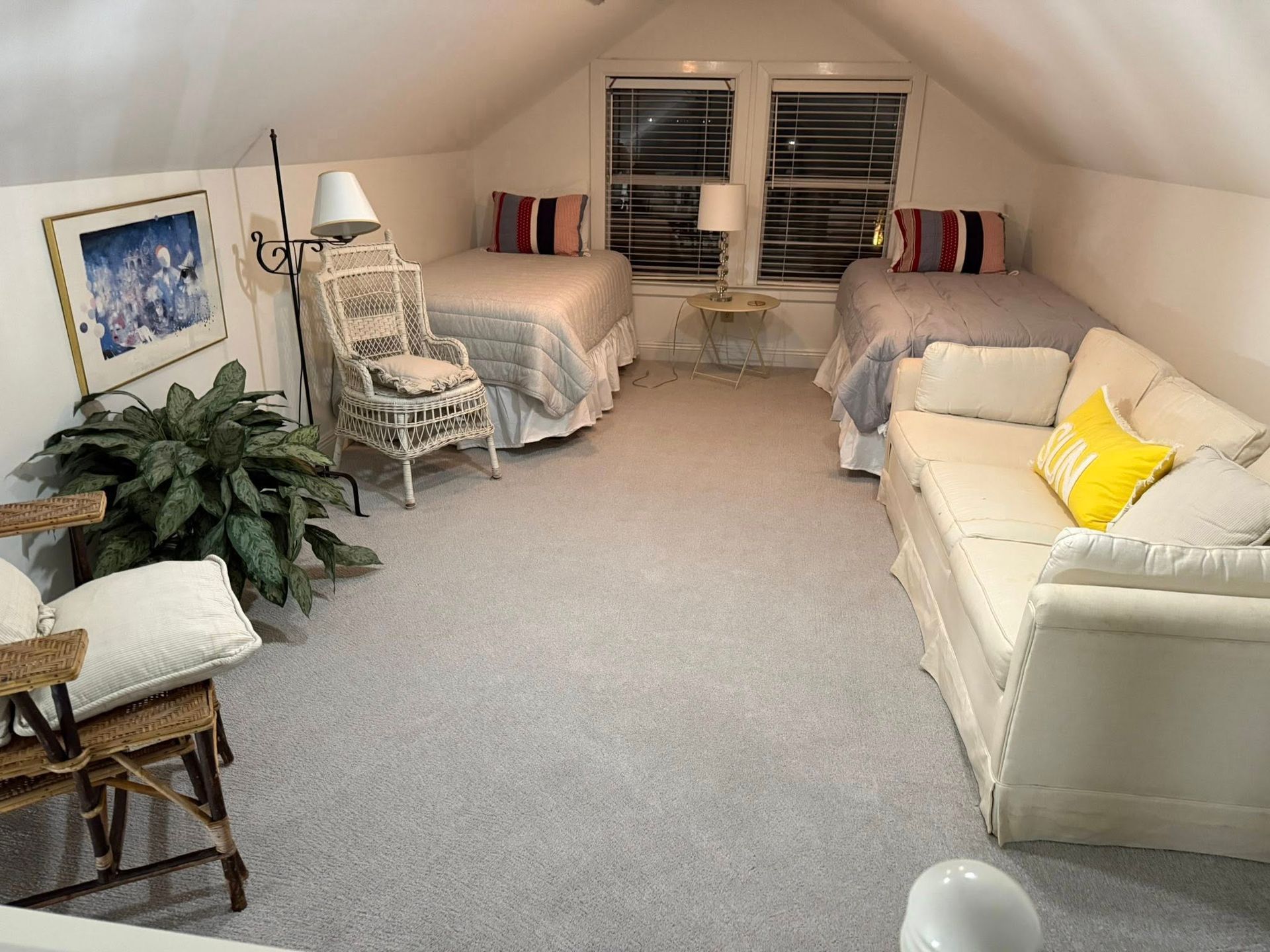 Attic room with two twin beds, a white sofa with a yellow pillow, a wicker chair, and a large plant.