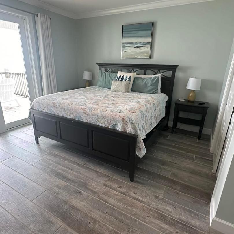A modern bedroom with a dark wooden bed frame, patterned bedding, two nightstands, and light gray wood-look tile floors.