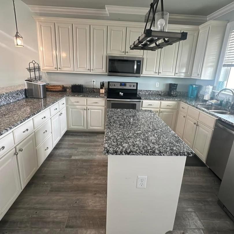 A well-lit kitchen with white cabinets, dark granite countertops, a center island, and gray wood-look plank flooring.