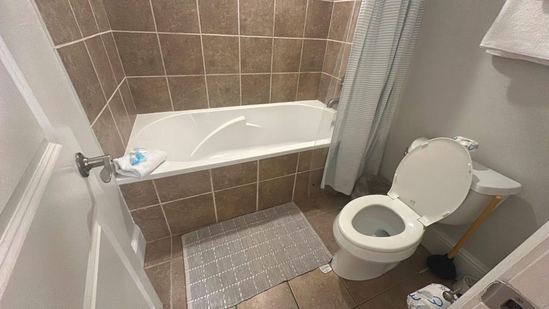 A small, neutral-toned bathroom with a tiled bathtub, shower curtain, and a white toilet with the lid open.