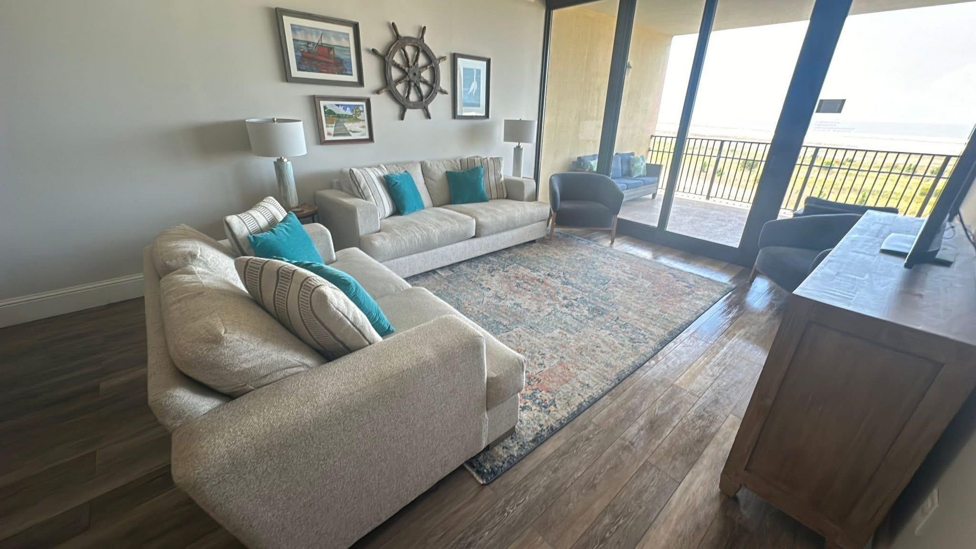 A living room with two neutral couches, turquoise accent pillows, nautical wall art, and a view of the beach outside.
