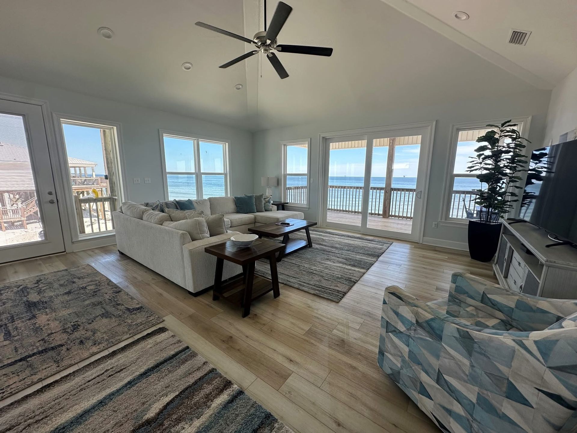 A bright living room with white walls, a sectional sofa, patterned armchair, and large windows overlooking the ocean.