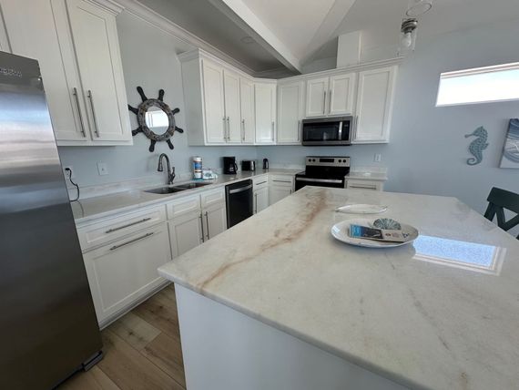 A bright kitchen with white cabinets, a large marble-topped island, stainless steel appliances, and beach-themed decor.