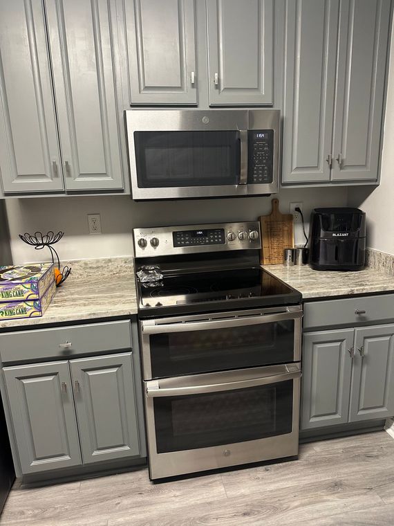 A modern kitchen featuring grey cabinets, granite countertops, a stainless steel microwave, double oven, and air fryer.
