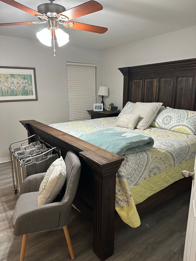 A bedroom featuring a large dark wooden bed with a yellow and green patterned quilt, a grey chair, and a ceiling fan.
