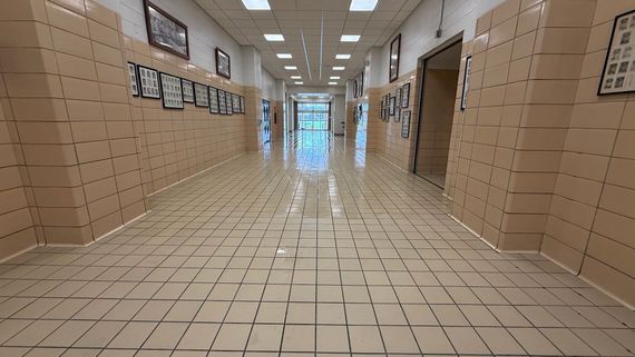 A long, empty school hallway with beige tiled walls and floors, illuminated by rows of ceiling lights.