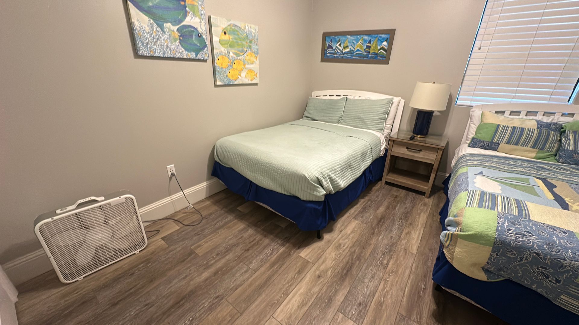 A bedroom with two beds, a nightstand with a lamp, a box fan on the floor, and nautical-themed wall art.