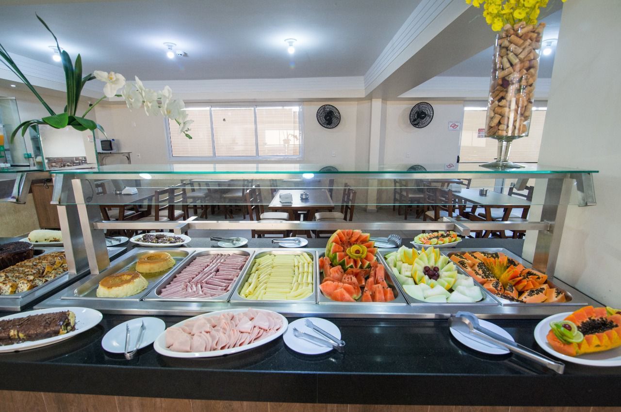 Buffet com comidas variadas, incluindo frutas, carnes e doces. Área de jantar bem iluminada.