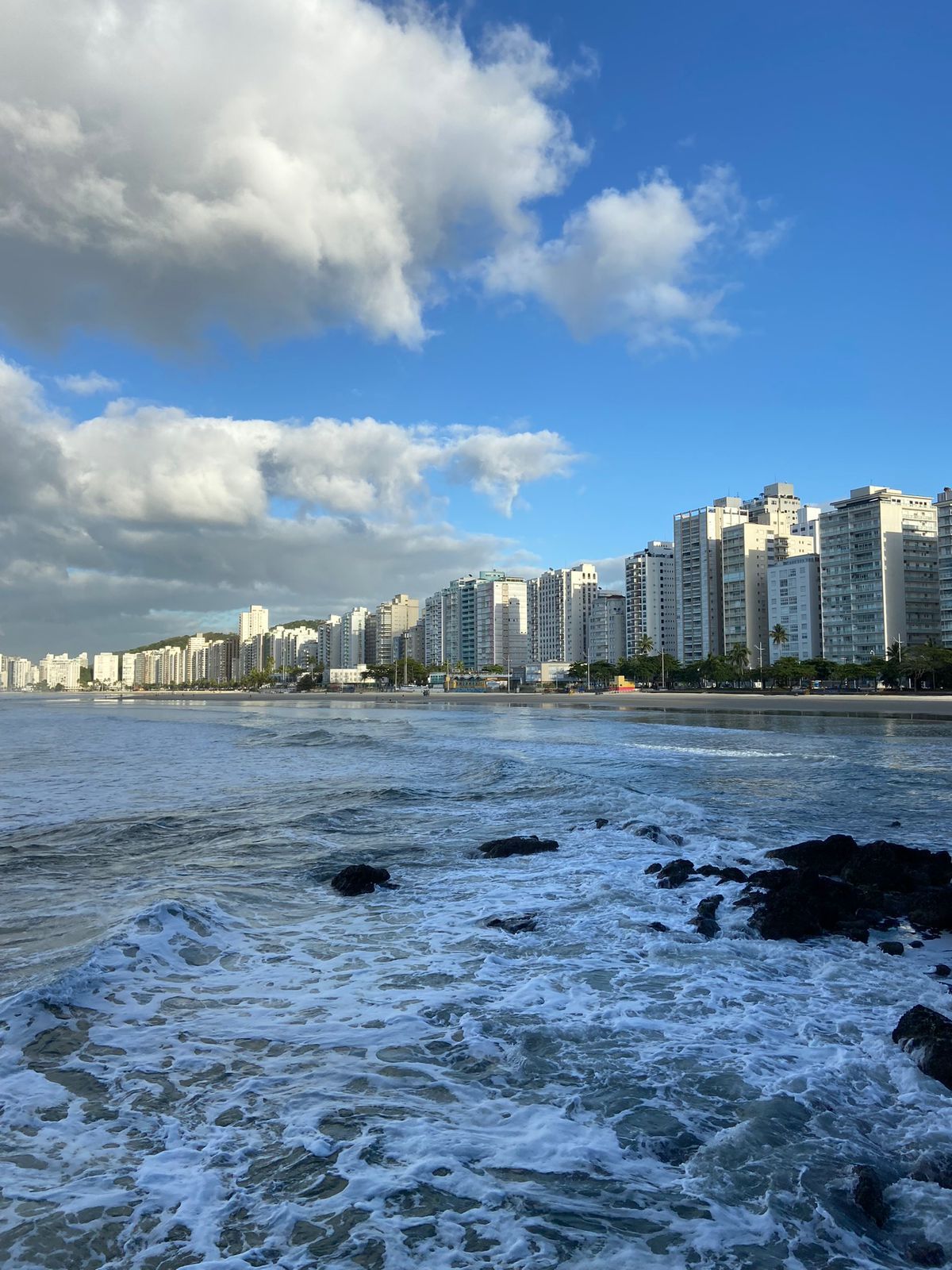Ondas do oceano quebrando em uma costa rochosa, com um horizonte de edifícios brancos sob um céu azul com nuvens fofas.