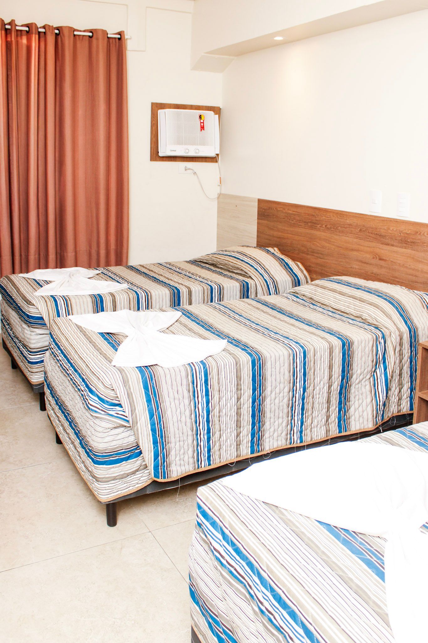 Duas camas de solteiro em um quarto de hotel com roupa de cama listrada de azul e branco, cortinas marrons e cabeceira.