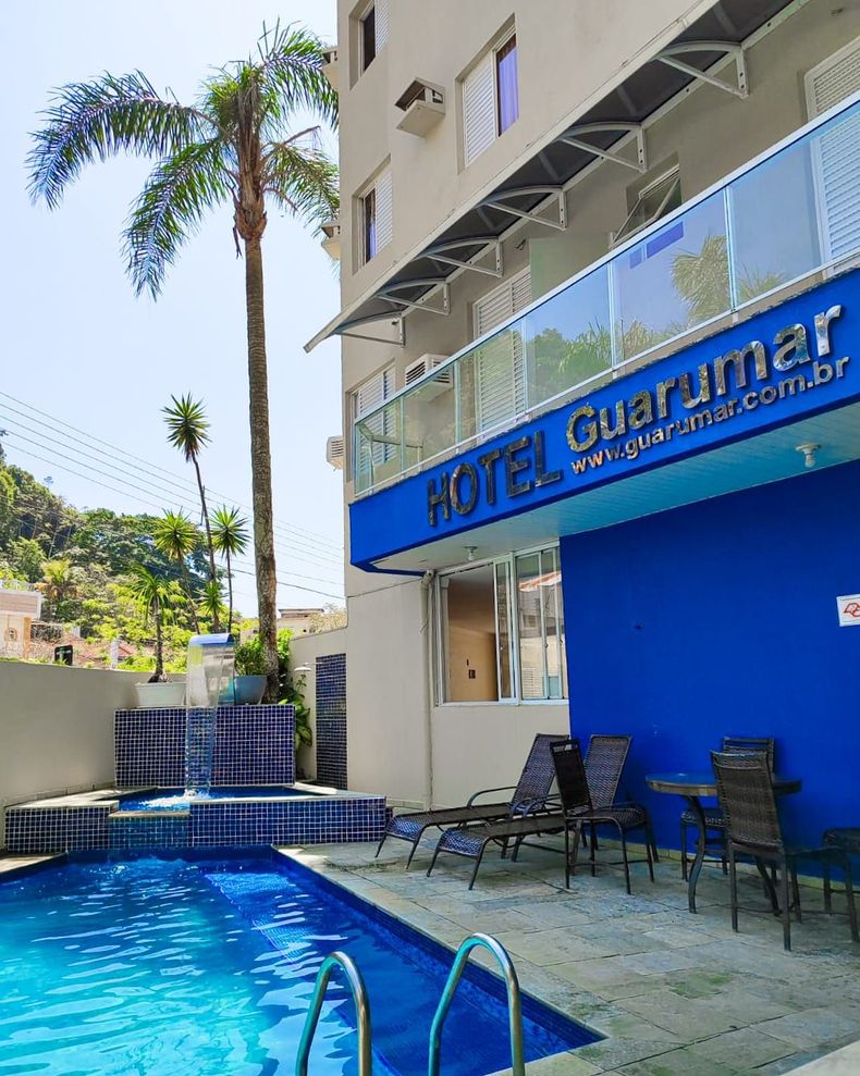 Hotel com fachada azul e piscina, dia ensolarado. Palmeira, cadeiras à beira da piscina, endereço do site visível.